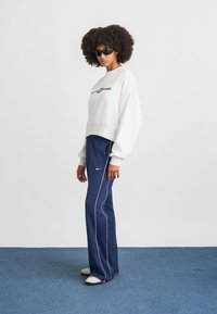 Szürke rövidített pulóver logóval, sötétkék sportnadrággal, fehér oldalsávokkal, és bézs slip-on cipővel párosítva; sötét napszemüveg viselve.