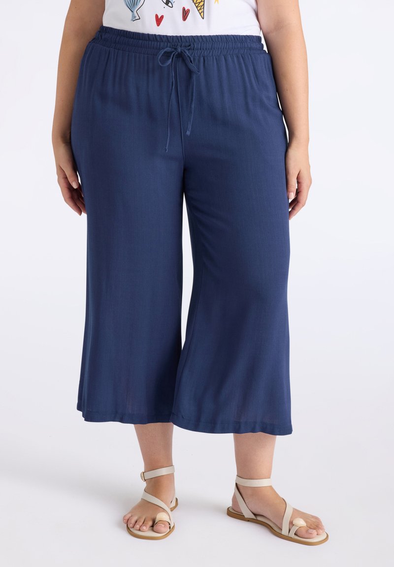 Pantalons larges bleu marine en matériau léger avec une taille élastique et un cordon de serrage. Associés à des sandales de couleur claire.