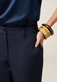 Persona vestita con pantaloni blu navy e una blusa in seta blu navy, con tre bracciali di grandi dimensioni beige e neri al polso, mano in tasca.
