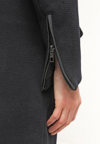 Black wool-blend coat detail showing a zipper on the cuff, with a smooth leather trim and a textured fabric finish.