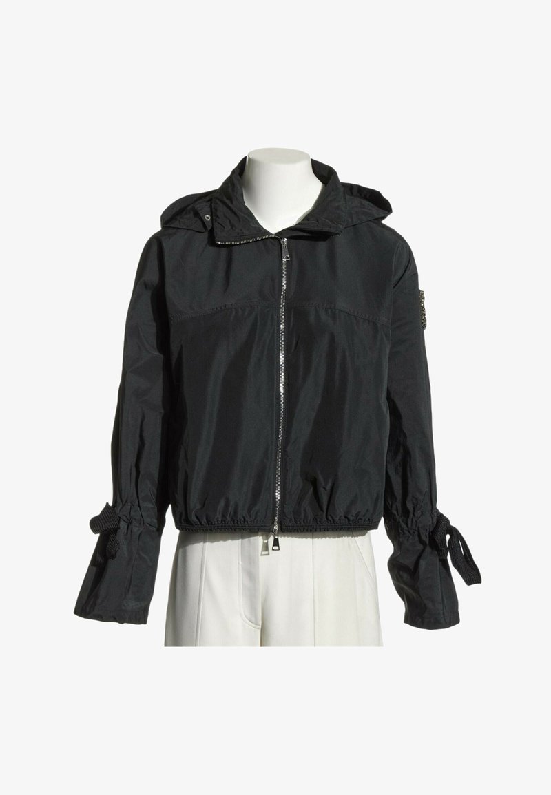 Veste à capuche noire zippée avec poignets froncés noués en nœuds, portée sur un mannequin blanc, associée à un pantalon blanc.
