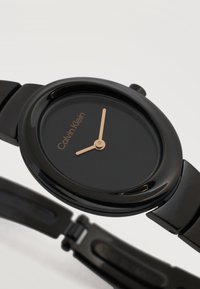 Μαύρο ρολόι χειρός Calvin Klein με χρυστά ωριαία και λεπτά χέρια σε στρογγυλό καντράν και μεταλλικό μπρασελέ με συνδεδεμένα κρίκους.