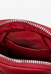Rotes Innenfutter einer Tasche mit glatter, satinähnlicher Textur und einer Ledertasche, die mit "Desigual" geprägt ist. Silberner metallischer Reißverschluss mit weißen Zähnen.
