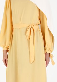Yellow dress with long puff sleeves and a tied waist belt, featuring a smooth texture and a draped design, creates an asymmetrical silhouette.