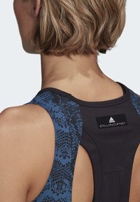 Černý atletický top s modrým hadím vzorem na ramenou, design s otevřenými zády a černá štítek s logy Adidas a Stella McCartney.