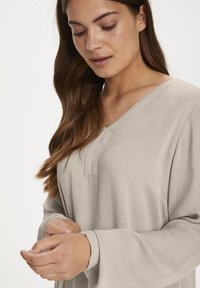 Világ bézs, hosszú ujjú blúz V-kivágással és gombos részletekkel. Puha, texturált anyag, laza szabással és felgöngyölt ujjakkal. Női modell hullámos hajjal.
