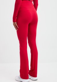 Czerwone legginsy w stylu dzwonów, wykonane z gładkiego materiału, z wysokim stanem i dopasowanym krojem, w połączeniu z białymi butami sportowymi.