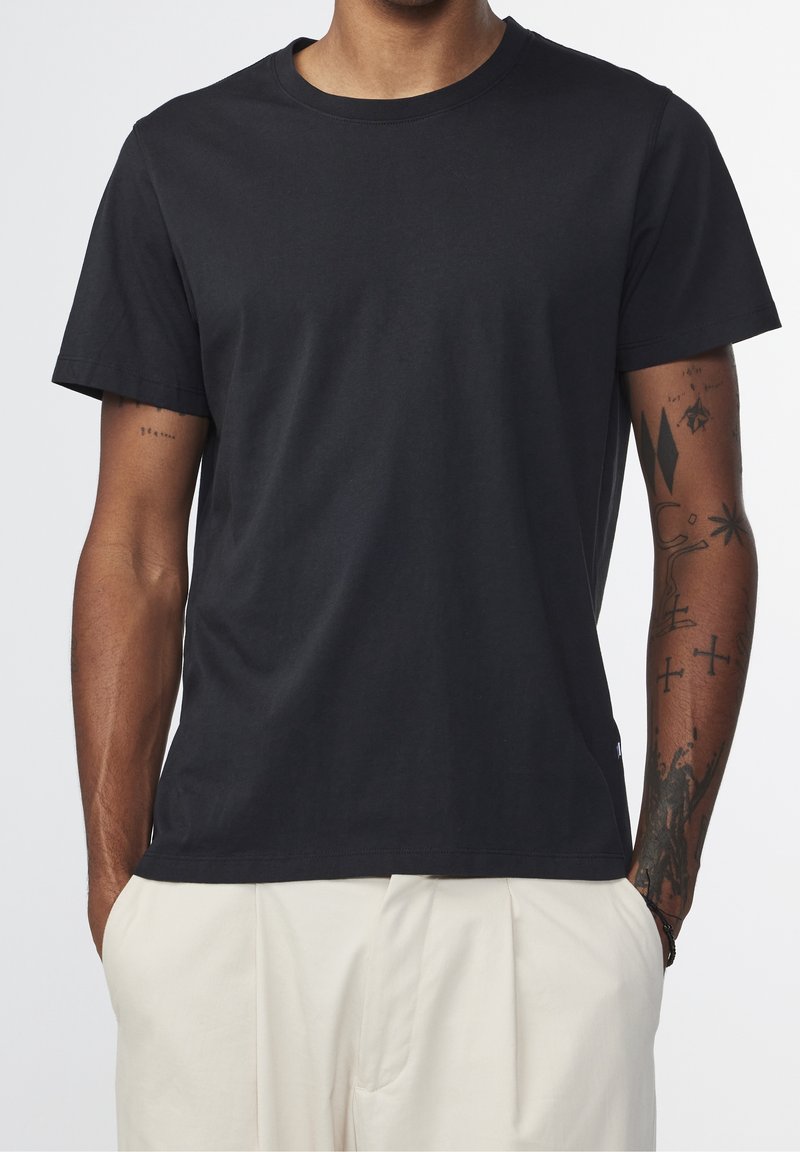 Homme portant un t-shirt noir uni à manches courtes et un pantalon beige clair, les bras tatoués, les mains dans les poches, sur un fond uni.
