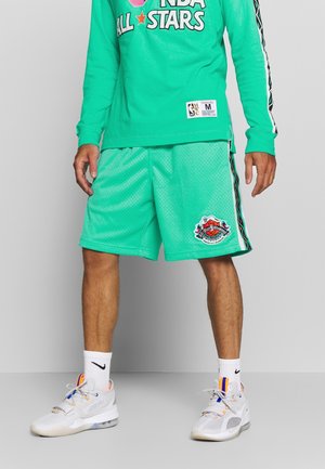 Homme portant un t-shirt à manches longues turquoise NBA All Stars et un short en mesh, avec des baskets Nike blanches et des chaussettes, sur fond gris.