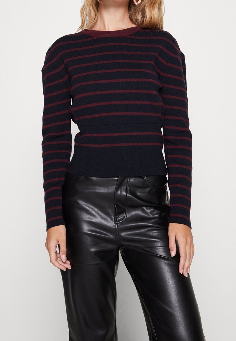 Femme portant un pull à manches longues rayé noir et rouge foncé associé à un pantalon en cuir noir taille haute, visible des épaules aux hanches.