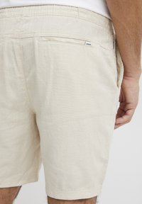 Beige rövidnadrág könnyű anyagból, laza fazonnal, rugalmas derékrésszel és egyetlen hátsó zsebbel, rajta címkével.