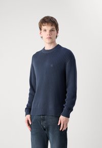 Maglione navy a coste con collo tondo, maniche lunghe e branding discreto sul petto. Il tessuto ha un aspetto strutturato.