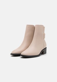 Béžové syntetické kotníkové boty s špičatou špičkou, nízkým blokovým podpatkem a zlatým akcentem na boku. Hladká textura a minimální šití.