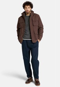 Giacca utility marrone con multiple tasche frontali, maglione grigio sotto, pantaloni blu navy e scarpe marroni, in piedi davanti a uno sfondo bianco.