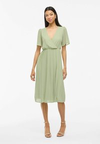 Femme portant une robe midi plissée vert clair avec des manches courtes évasées et un décolleté en V profond, assortie à des sandales à talons dorées ouvertes.