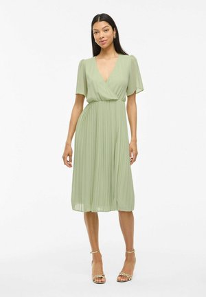 Femme portant une robe midi plissée vert clair avec des manches courtes évasées et un décolleté en V profond, assortie à des sandales à talons dorées ouvertes.