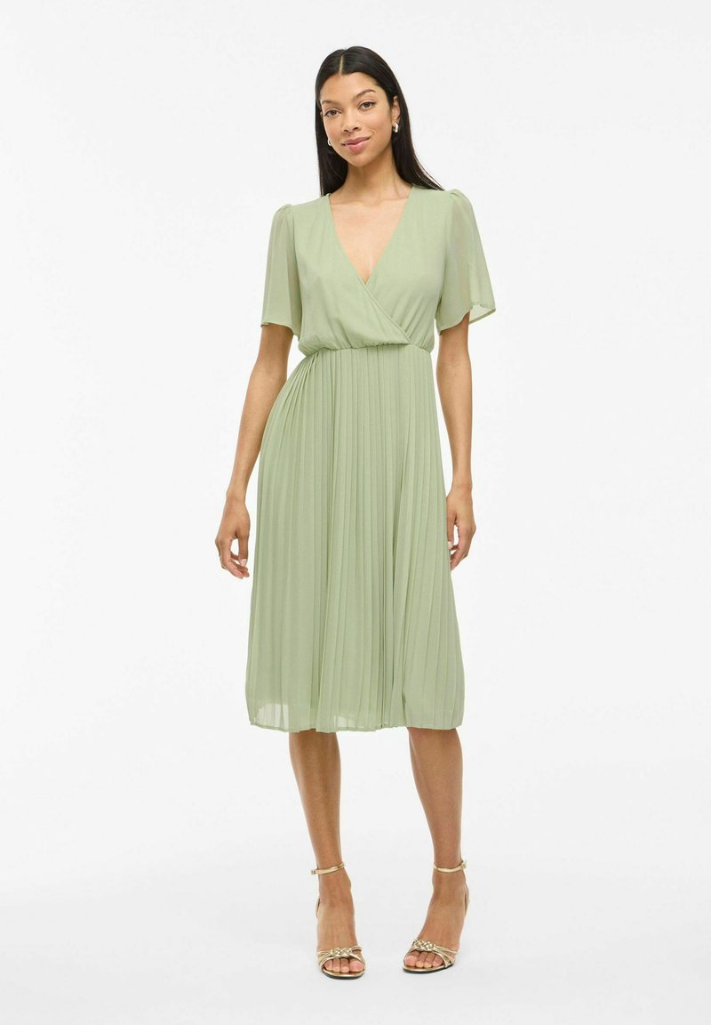 Femme portant une robe midi plissée vert clair avec des manches courtes évasées et un décolleté en V profond, assortie à des sandales à talons dorées ouvertes.