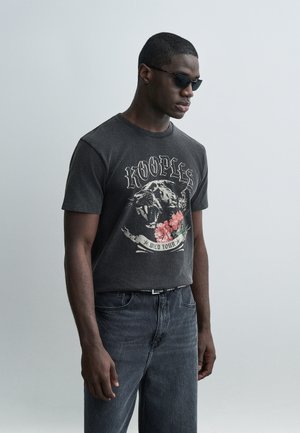 Jeune homme portant des lunettes de soleil noires, un t-shirt graphique noir avec une panthère rugissante et des fleurs, associé à un jean foncé sur un fond uni.