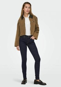 Giacca corta marrone con colletto nero, maglia bianca a coste, jeans skinny blu scuro e mocassini neri. Design semplice, vestibilità casual.