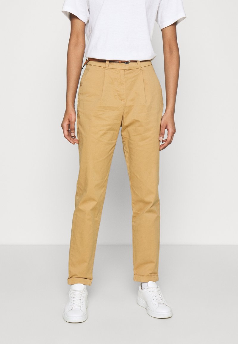 Pantalon en coton couleur fauve à coupe fuselée, avec des plis sur le devant et des poches latérales, assorti de baskets blanches et d'un t-shirt blanc uni.