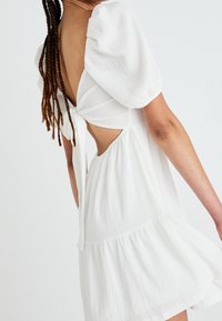 Weißes strukturiertes Kleid mit Puffärmeln, offenem Rücken mit Bindeband und ausgestelltem Rock mit Volantabschluss.