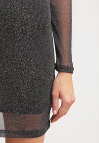 Musta kiiltävä mekko, jossa on teksturoitu kangas ja kimaltava kuvio. Pitkät läpinäkyvät hihat ja vartalonmyötäinen muotoilu.