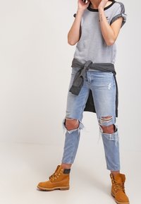 Grijs shirt met korte mouwen en een zwarte halslijn; blauwe gescheurde spijkerbroek met rafelige zoom; bruine veterlaarzen met een gestructureerd bovenwerk en een rubberen zool.