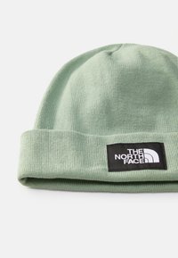 Beanie lavorato a maglia verde chiaro con risvolto, dotato di un'etichetta nera con testo e logo bianchi. Texture morbida e design semplice.