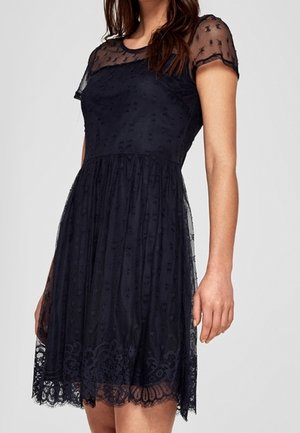 Femme portant une robe noire en dentelle longueur genou avec des manches courtes transparentes et des détails brodés floraux sur la jupe et le corsage.