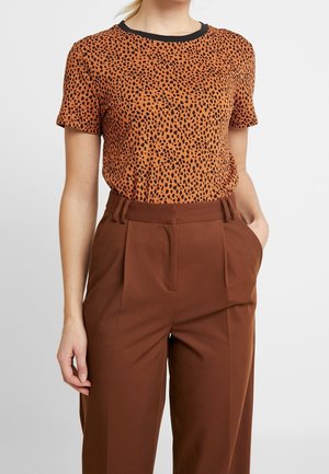 Persoon draagt een oranje-zwart gevlekt shirt met korte mouwen, ingestopt in een hoog getailleerde bruine geplooide broek, met handen in de zakken.