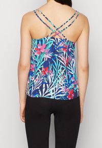 Femme portant un haut sans manches à motifs floraux avec des bretelles fines croisées et un legging noir, debout de dos sur un fond neutre.