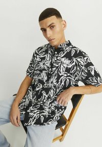 Camisa negra de manga corta con estampado floral blanco, cuello abotonado y corte relajado. Llevada con pantalones de mezclilla azul claro.