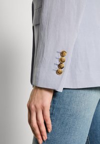 Jasnoniebieska marynarka o fakturowanej powierzchni, z mankietem ozdobionym czterema guzikami. Noszona na dopasowanych dżinsach, prezentująca krojony fason.