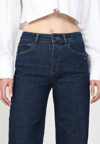 Dunkelblaue Jeans mit hoher Taille, klassischem Fünf-Taschen-Design und einem Markenlabel an der Seite, glatte Textur.