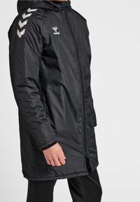 Man som bär en svart knälång huvmantel med blixtlås och vita chevronformade logotyper på bröstet och axeln.