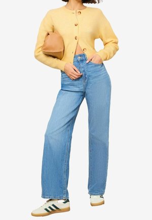 Femme portant un cardigan jaune boutonné, un jean bleu taille haute à jambes larges, des baskets blanches avec des rayures vertes, tenant un sac à main beige.