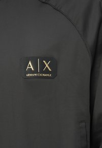 Μαύρο ύφασμα με ραμμένο μαύρο πατς που εμφανίζει χρυσά γράμματα "A|X" και το λογότυπο "Armani Exchange".