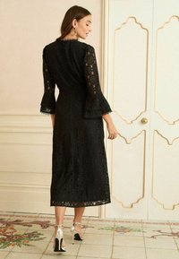 Must mustriga kleit pika kellukese varrukaga, millel on keeruline lillemuster. Kleit ulatub põlvedeni ja on sobitatud hõbedaste kõrgete kontsadega.