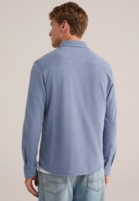 Muž v světle modré košili s dlouhým rukávem a světlých džínách stojí zády k nám před jednobarevným pozadím.