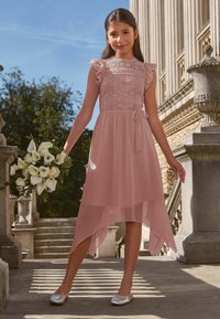Világos rózsaszín ruha csipkebetéttel az elején, fodros ujjal és aszimmetrikus szegéllyel. Ezüst lapos cipővel párosítva, fehér virágcsokrot tartva.