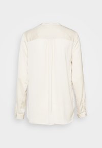 Camicia cremosa a maniche lunghe con yoke e dettaglio arricciato nella parte superiore della schiena, con polsini bottonati, esposta su uno sfondo chiaro e semplice.