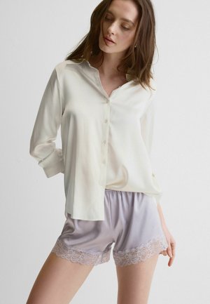 Frau trägt eine cremefarbene Seidenbluse mit Knöpfen und lavendelfarbene Satinhosen mit Spitzenabschluss, steht vor einer schlichten weißen Wand.