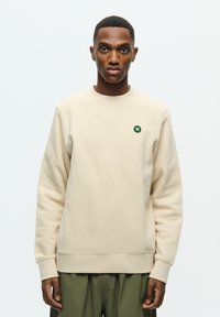 Cream sweatshirt made from soft material, featuring a round neckline and ribbed cuffs. A small green logo is positioned on the chest.
