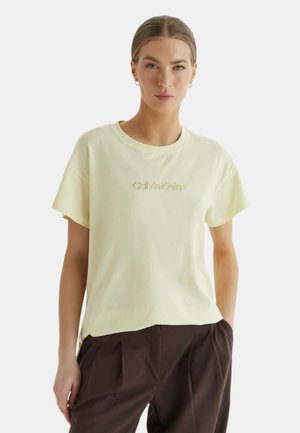 Donna con capelli castano chiaro raccolti in uno chignon basso, che indossa una maglietta giallo pallido Calvin Klein e pantaloni plissettati marrone scuro con le mani nelle tasche.