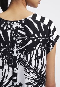 Camicia bianca e nera con una stampa botanica sul corpo e audaci strisce sulle spalle, con chiusure a bottoni blu sul retro.