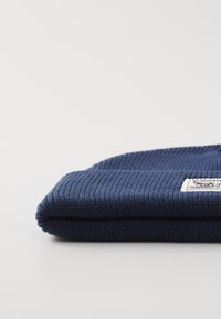 Navy blue knit beanie with ribbed texture, folded over at the top. Features a small label on the side with text and graphic.