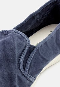 Morska platnena obutev za slip-on z elastičnimi stranskimi paneli in belim gumijastim podplatom. Teksturovana zgornja tkanina se ponaša s subtilnim leskom in vidnimi šivi.