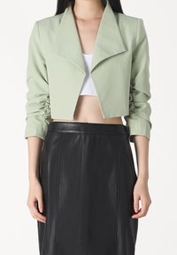 Veste courte en tissu vert clair avec un large col et des manches froncées. Associée à une jupe en cuir noir, mettant en valeur une texture lisse.