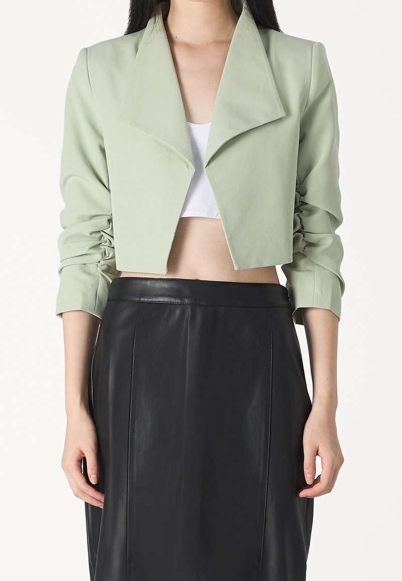 Veste courte en tissu vert clair avec un large col et des manches froncées. Associée à une jupe en cuir noir, mettant en valeur une texture lisse.