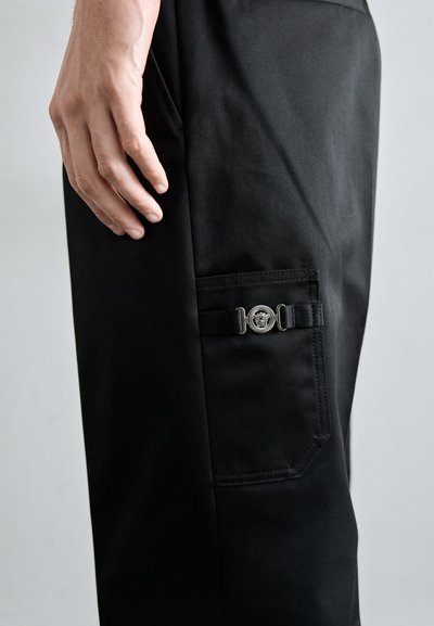 Black pants made of smooth fabric, featuring a side pocket with a metallic button closure and a subtle sheen. Tailored fit, no patterns.